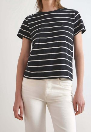 Personne portant un haut à manches courtes rayé noir et blanc à rayures horizontales et un pantalon blanc taille haute, se tenant devant un fond clair.