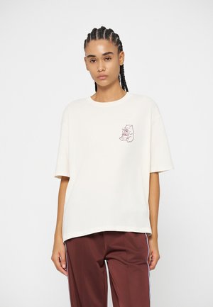 Femme aux cheveux tressés portant un T-shirt blanc oversize avec un petit motif d'ours et un pantalon marron avec des bandes latérales.