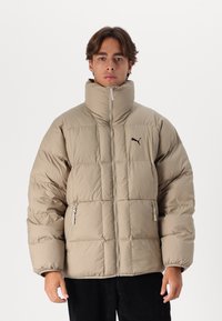 Il giubbotto trapuntato beige presenta un colletto alto, una texture a righe orizzontali, due cerniere laterali e un logo nero nella zona sinistra del petto.