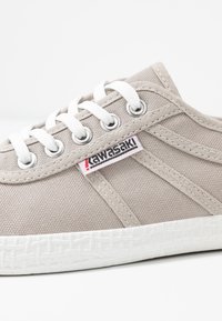 Beige canvas sneaker with white laces, metal eyelets, textured fabric, and a white rubber sole featuring a patterned design, branded label visible.