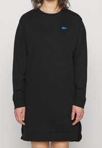 Vestido negro de manga larga hecho de una tela suave, con un cuello redondeado, aberturas laterales y un pequeño logo azul en el pecho.