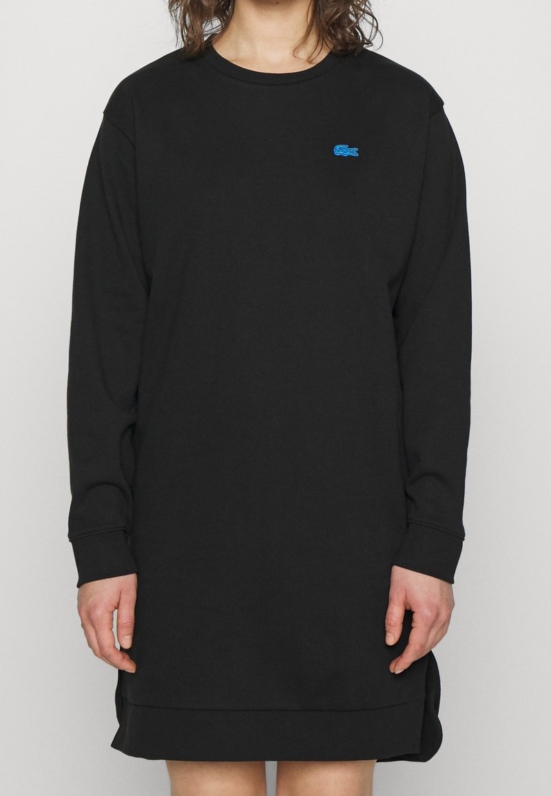 Vestido negro de manga larga hecho de una tela suave, con un cuello redondeado, aberturas laterales y un pequeño logo azul en el pecho.