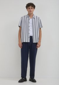 Chemise à manches courtes avec un motif géométrique en bleu marine et blanc, associée à un pantalon bleu marine et des chaussures noires, le tout sur un fond neutre.