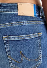 Denim jeans with a worn blue texture, featuring contrasting yellow stitching, a leather label at the waistband, and a back pocket design.