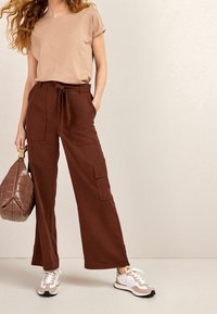 Beige short-sleeve top and brown wide-leg trousers with a tie waist. Brown quilted handbag and light-colored sneakers with accents.