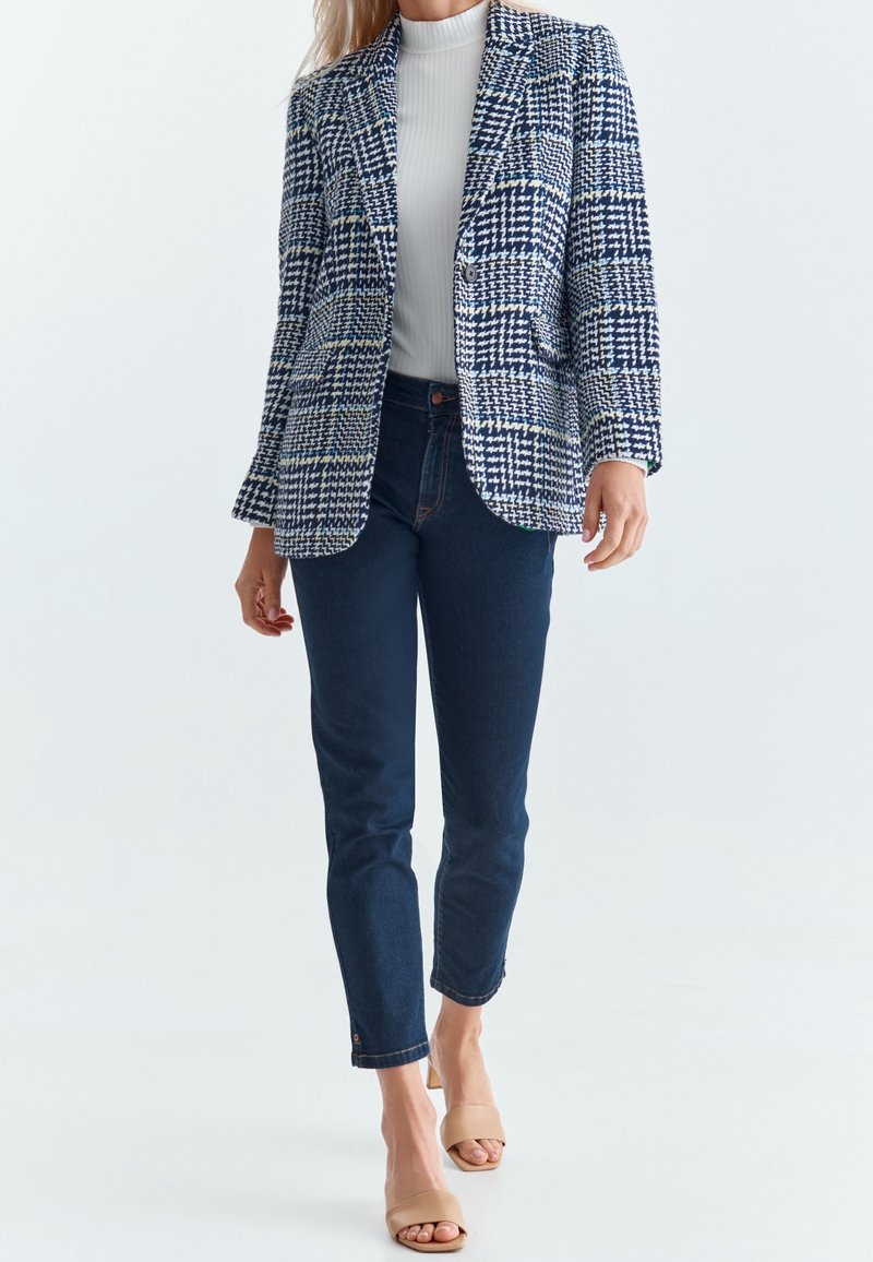 Femme portant un blazer à motif pied-de-poule bleu et blanc, un col roulé blanc côtelé, un jean skinny foncé et des talons ouverts beige.
