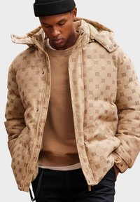 Beige prošívaná bunda s vzorovaným designem, zipem a patentkami, předními kapsami a kapucí. Nalezená na hnědém svršku.