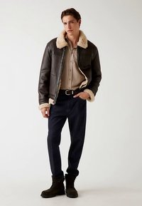 Braune Lederjacke mit Shearling-Kragen, beige Hemd mit Knöpfen, dunkle Jeans und schwarze Stiefel. Modell steht vor neutralem Hintergrund.
