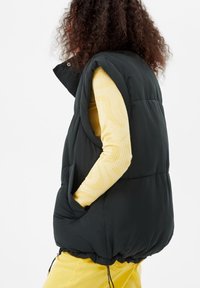 Black puffer vest with a high collar and side pockets, worn over a yellow ribbed top. Features a drawstring hem and textured fabric.