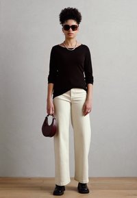 Haut noir à manches longues, pantalon large crème, bottes foncées et petit sac à main bordeaux. Le mannequin porte des lunettes de soleil surdimensionnées et un collier.