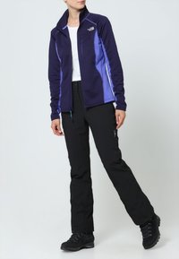 Polarowa kurtka w ciemnofioletowym kolorze z jasnofioletowymi akcentami, z kieszenią na zamek i wysokim kołnierzem; zestawiona z czarnymi spodniami i solidnymi butami.