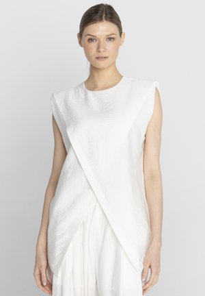 Młoda kobieta z zaczesanymi do tyłu włosami nosi bezrękawnik biały top w stylu kopertowym i dopasowane białe spodnie na jednolitym jasnym tle.