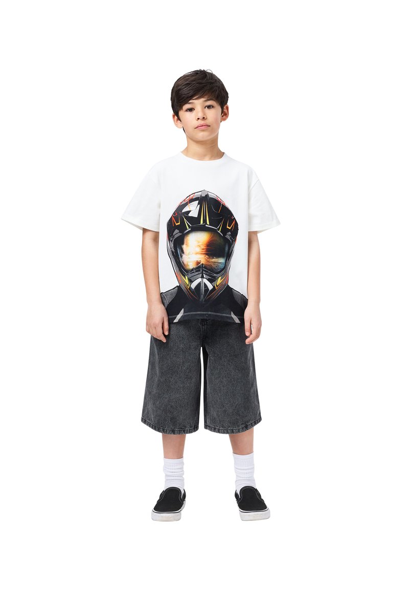 Λευκό T-shirt κοντό μανίκι με γραφικό σχέδιο ενός κράνους. Συνδυασμένο με σκούρες γκρι φαρδιές βερμούδες και μαύρα slip-on παπούτσια με λευκές κάλτσες.