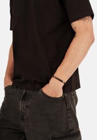 Schwarzes Baumwoll-T-Shirt mit kurzen Ärmeln, kombiniert mit dunkelgrauen Jeans. Ein dünnes, strukturiertes Armband wird am Handgelenk getragen.