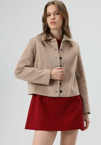 Beige, cropped jack in zachte stof met een knoopsluiting aan de voorkant en een spitse kraag. Gecombineerd met een rode jurk, wat zorgt voor een contrasterende uitstraling.