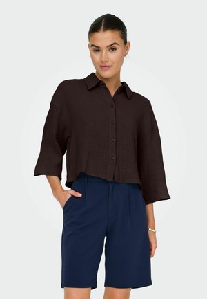 Chemise courte marron à boutons en tissu texturé avec un col, associée à un short plissé bleu marine. Le modèle se tient avec une main dans la poche.