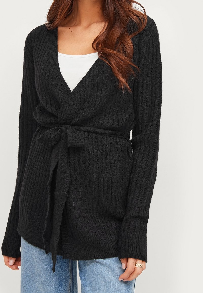 Schwarzer, gerippter Cardigan mit V-Ausschnitt und Bindegürtel in der Taille. Lange Ärmel und weiche Textur, kombiniert mit einem weißen Top darunter.