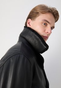 Mustat nahkatakki korkealla kauluksella, jossa on mustaa turkista vuori, hieno rakenne ja sileä pinta, kuvattuna sivulta.