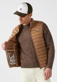 Gilet imbottito marrone con trapuntatura verticale, maglione beige sotto e berretto bicolore con logo. Stampe interne visibili nel gilet.