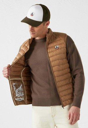 Brown puffer vest with vertical quilting, tan sweater underneath, and a two-tone cap featuring logo. Interior prints visible in vest.