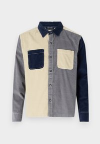 Knappeskjorte med et lappeteppe-design av grå, marineblå og beige kordfløyelsstoff. To frontlommer, en i marineblå og en i beige.