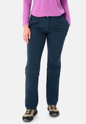 Person, die dunkelblaue Wanderhosen mit Reißverschluss, beige Wanderschuhe mit schwarzen Sohlen und ein langärmliges lila Oberteil trägt, steht mit einer Hand in der Tasche.