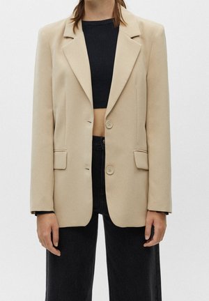 Blazer beige sur mesure à deux boutons porté sur un top court en maille côtelée noir et un pantalon noir taille haute.