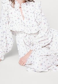 Valkoinen kukallinen mekko, jossa on röyhelökoristeita, sinisiä ja vaaleanpunaisia kukkia. Pitkät hihat ja solmittava kaula-aukko, valmistettu kevyestä kankaasta.