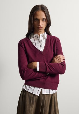 Mujer joven con cabello largo que lleva un suéter burdeos con cuello en V sobre una camisa blanca de rayas y una falda plisada marrón, con los brazos cruzados.