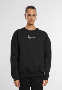 Felpa nera realizzata in morbido tessuto, con colletto a girocollo, vestibilità oversize e logo bianco sul petto. Polsini e orlo a costine.
