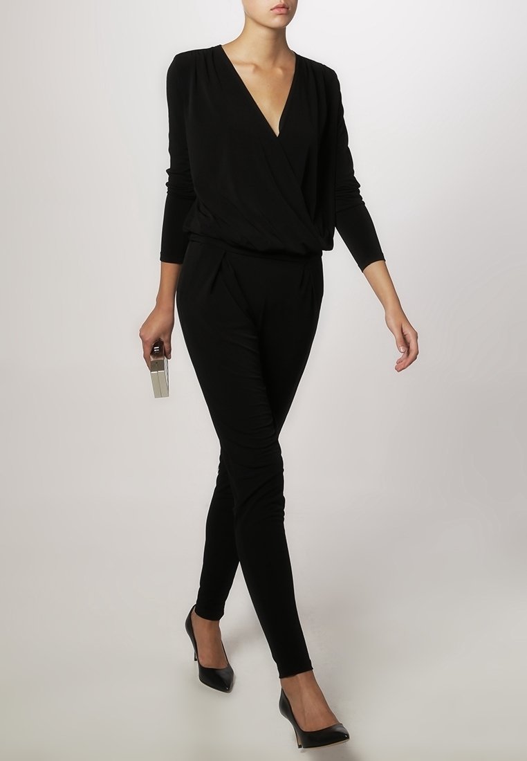 Mono negro con mangas largas, escote en V, corte a medida, cintura elástica y piernas ajustadas. Llevado con tacones negros de punta y sosteniendo un clutch.