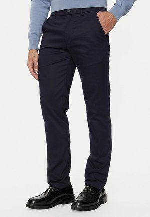 Pantalons de costume bleu marine en tissu lisse, présentant une coupe classique, des poches latérales et une fermeture à bouton. Associés à des chaussures noires brillantes.