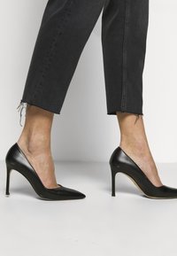 Zapatos de tacón alto de cuero negro con punta afilada y tacón de aguja. Usados con jeans negros recortados con dobladillos deshilachados.