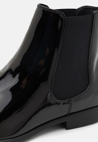 Bottine en cuir verni noir avec des panneaux latéraux en élastique, bout arrondi et petit talon empilé. Surface lisse avec peu de détails de couture.