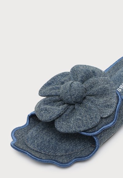 Pantoufle en denim avec une grande fleur rembourrée en denim sur le dessus et un bord festonné, présentée sur un fond blanc uni.