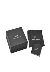 Sort rektangulær æske med et hvidt "A/X ARMANI EXCHANGE" logo, ledsaget af en sort pose og et lille hæfte, alt i matchende farver.