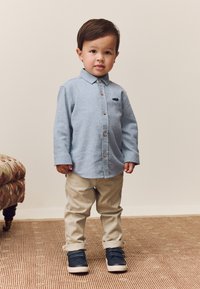 Camicia azzurro chiaro con bottoni e piccolo logo, pantaloni beige e sneaker blu navy. La camicia ha maniche lunghe e bottoni in legno.