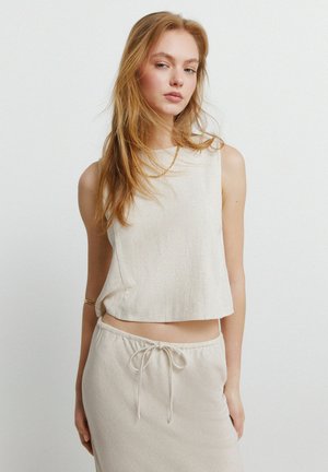 Ung kvinde med langt rødt hår iført en ærmeløs beige top, matchende nederdel med snøre, guldkæde og armbånd mod en ensfarvet baggrund.