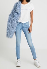 Ljusblå skinny jeans i kombination med en vit t-shirt med texten "INFLUENCER" i pastellfärger. Ett fluffigt ljusblått plagg hänger över ena axeln.