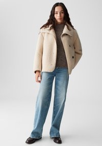 Beige Kunstfellmantel mit einem Schalkragen und schwarzen Knöpfen, kombiniert mit einem grauen Pullover, hellblauen weit geschnittenen Jeans und dunkelbraunen Schuhen.