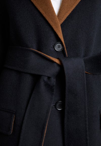 Wollmantel in Marineblau mit einem braunen Kragen. Verfügt über einen Gürtel, zwei Vordertaschen und zwei schwarze Knöpfe. Weicher Stoff mit tailliertem Schnitt.