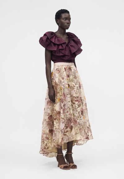 Γυναίκα με μπορντό τοπ με βολάν και φούστα με φλοράλ σχέδιο, με ψηλές μπεζ σανδάλια, που στέκεται σε απλό ανοιχτό φόντο.