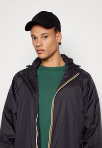 Veste légère noire avec capuche, dotée d'une fermeture éclair multicolore et d'un t-shirt vert en dessous. Une casquette noire complète la tenue.