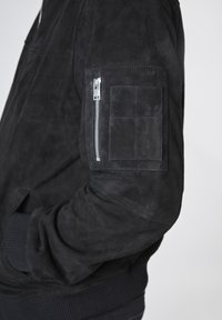 Svart mockabomberjacka med ribbstickade ärmar, har en dragkedjeficka på vänstra armen och en jämn textur.