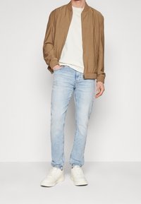 Brun bomberjacka, vit T-shirt, ljusblå jeans och vita sneakers. Jackan har ribbad mudd och krage; jeansen har slim-fit.