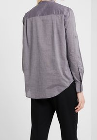 Camisa de manga larga gris con un panel trasero en un tono gris más claro, con detalle de puño con botón y un ajuste relajado. Tejido suave y texturizado.