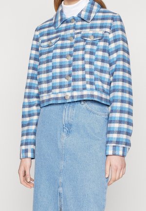 Kobieta w niebiesko-białej, kraciastej, krótkiej kurtce z srebrnymi guzikami, połączonej z jasnoniebieską jeansową spódnicą i białym golfem.