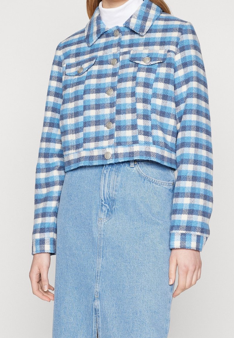 Femme portant une veste courte à carreaux bleue et blanche avec des boutons argentés, assortie d'une jupe en jean bleu clair et d'un col roulé blanc.
