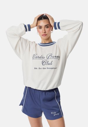 Kvinde med mørkt hår i hvid sweatshirt og blå shorts, stående med hænderne på hovedet mod en ensfarvet hvid baggrund.