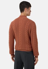 Pull en tricot couleur rouille avec motif à torsades, poignets côtelés et col montant. Présente une coupe décontractée et une surface texturée.